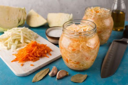 Open glass jars of sauerkraut on blue background. Next to is sliced fresh cabbage and carrots, bay leaf and garlic. Homemade salad. Fermented food.の写真素材