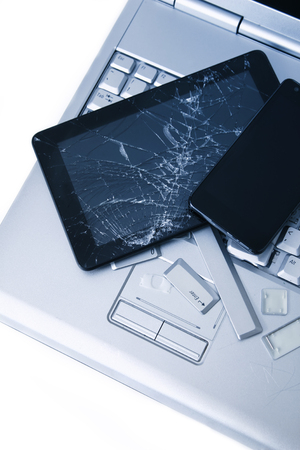 A silver laptop with a broken keyboard, tablet with a cracked display and black phone. A close-up picture of part of broken laptop and cracked screen on a tablet.の写真素材