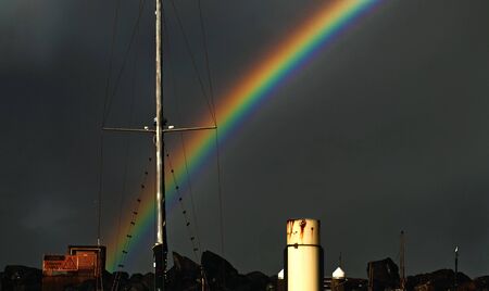 Colourful vivid waterfront rainbow with yacht masts and mooring pylon against the backdrop of a dark sky.Coffs Harbour, New South Wales, Australia.の写真素材