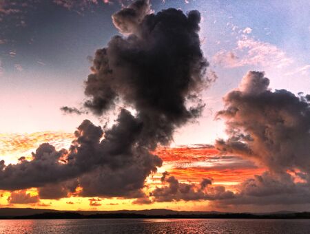 A striking vivid threatening charcoal and red coloured cloudy tropical sunrise seascape featuring cloud silhouettes with ocean water reflections. Queensland, Australia.の写真素材