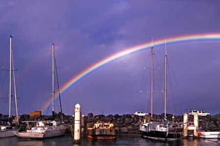 Rainbow in a dark blue sky over waterfront maritime marina/dock with berthed boats against a rock breakwall. Coffs Harbour, Australia.の写真素材