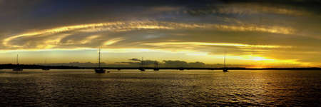 A striking inspirational golden yellow coloured cirrostratus cloudy coastal nautical Sunset Sesacsape in a blue sky over boats in sea water with water reflections. Tin Can Bay, Queensland, Australia.の写真素材