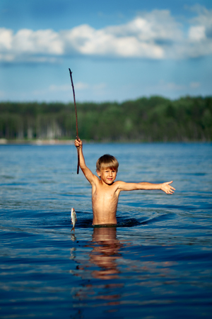 Little boy is fishing at the lake on a summer day with homemade fishing pole. Happy child catch a fish. Cute fisherman.の写真素材