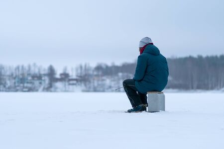 Fisherman on a frozen snowy lake. Back view. Winter fishing with ice hole. Man holding fishing rod in hands. Forest in the snow on the horizon.の写真素材