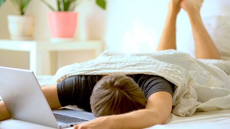 A man works in the bedroom at a laptop at home. Concept for remote work at home during the coronavirus quarantine period Covid 19.の写真素材