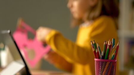 Distance education of children at home. A schoolgirl performs a creative task out of paper. In the foreground is a glass with colored pencils.の写真素材