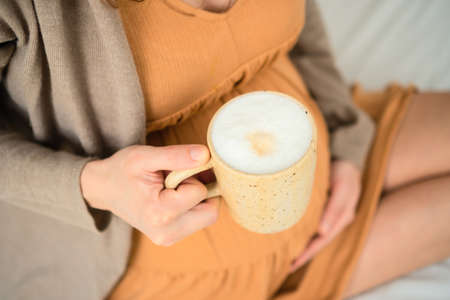 A cute pregnant woman holds in hand a cup of coffee. View from above. The concept of a happy pregnancy in anticipation of a baby. Not healthy drinks. 9 months of waiting for a baby.の写真素材