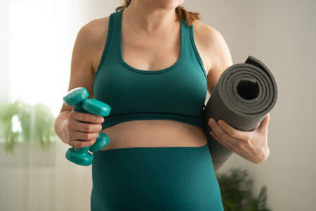 A pregnant woman prepare for exercising with dumbbells. Young girl holding a yoga mat in hand. Concept of a healthy lifestyle during pregnancy. Sports activities before the birth of baby.の写真素材