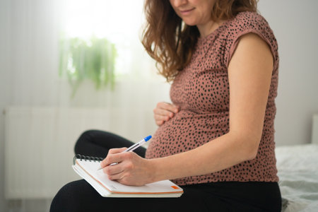 Pregnant woman making to-do lists in a notebook. Calm moment of preparation at home.の写真素材