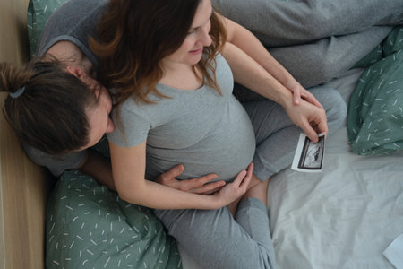 Aerial view shows expecting mother in lotus pose holding ultrasound scan while being embraced by her partner. Beautiful family anticipation scene.の写真素材