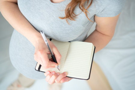 Future mother writing detailed maternity plan in notebook. Birds-eye close-up shows careful preparation of hospital checklist and birth reminders.の写真素材