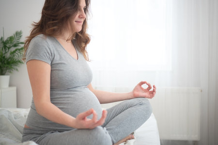 Serene expecting mother sitting cross-legged on bed, gently caressing her baby bump. Peaceful moment of maternal connection in cozy home setting.の写真素材