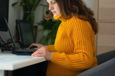 Close-up of expecting mother working remotely, showing pregnancy fatigue while managing workload. Warm natural light creates cozy yet professional atmosphere.の写真素材
