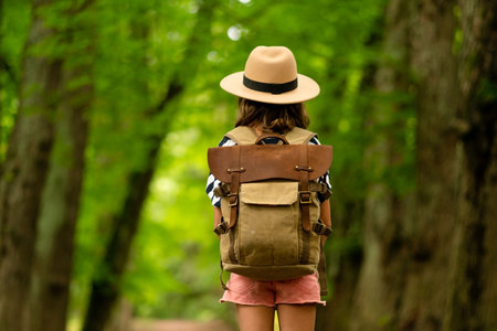 Child with hiking backpack stands at forest trailhead, back turned, poised for summer nature adventure. Symbol of youthful outdoor enthusiasm.の写真素材