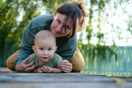 Mother and child sitting on a wooden surface in a garden setting, surrounded by greenery. The mother...