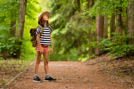 Curious young girl standing in sunlit forest during hiking trip. Backpack ready for summer nature exploration and outdoor fun.の写真素材