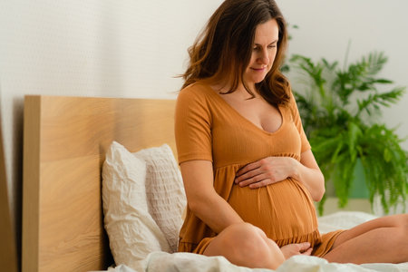Content expecting mother sitting on bed while gently caressing her pregnant belly. Intimate moment...