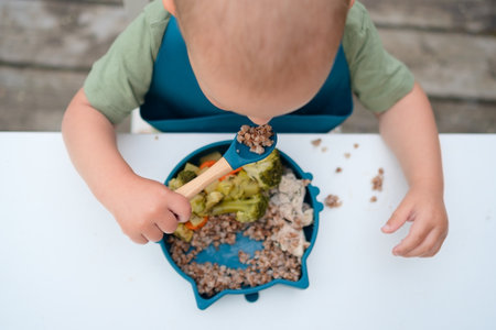 Top view of a toddler feeding themselves with a spoon outdoors. Healthy first meal with vegetables, sunny day.の写真素材