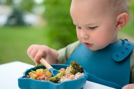 Toddler eating with a spoon from a bowl outdoors. Healthy baby meal with vegetables on a sunny day.の写真素材