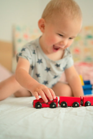 Curious baby playing independently with wooden train set on soft bed. Natural learning through tactile play in nurturing home setting.の写真素材