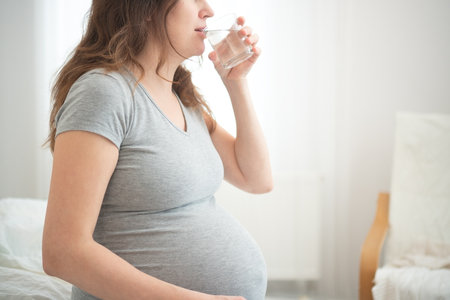 Expecting mother taking mindful sip from glass, maintaining hydration during pregnancy. Soft natural light enhances this health-conscious moment at home.の写真素材
