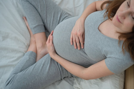 Radiant pregnant woman sitting on bed, lovingly touching her belly in cozy domestic setting. Beautiful anticipation of motherhood and new life.の写真素材