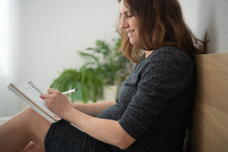 Pregnant woman writing a checklist in a notebook. Quiet moment of planning on bed.