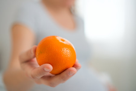 Expecting mother holding fresh orange while gently caressing her baby bump. Peaceful moment of healthy nutrition and maternal connection at home.の写真素材