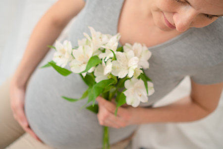 Expecting mother gently holding white flowers while touching her baby bump. Soft natural light enhances this tender maternal moment at home.の写真素材