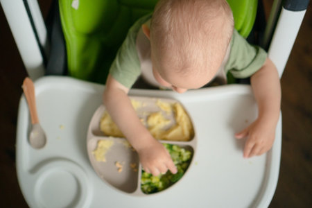 Top-down shot shows curious toddler in feeding chair picking nutritious foods - corn cereal, steamed veggies and egg. Important early eating skills development. Focus on child headの写真素材