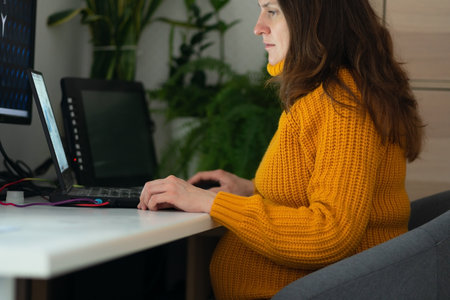 Expecting mother focused on laptop work, balancing pregnancy and career. Natural light highlights her tired but determined expression while managing professional duties.の写真素材