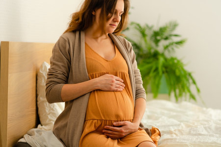 Radiant pregnant woman sitting on bed, lovingly touching her belly in cozy domestic setting....