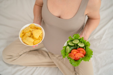 Pregnant woman holding both wholesome salad and salty chips. Authentic snapshot of dietary...