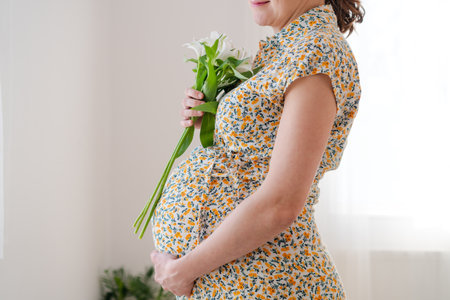 Expecting mother gently holding white flowers while touching her baby bump. Soft natural light...