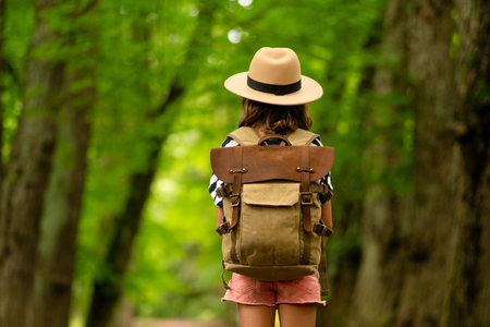 Young hiker stands ready in sunlit woods, backpack secured, beginning her summer adventure. Back...