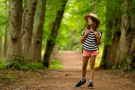 A full-body view of a dreamy girl walking with a backpack and hat, observing the forest around her. Peaceful summer hiking trip full of curiosity, nature, and exploration.の写真素材