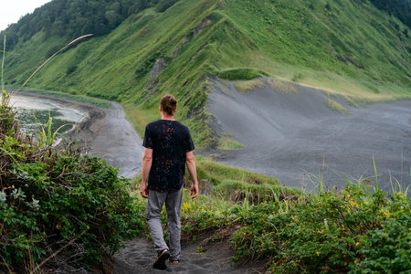 Solitary traveler standing on sandy beach with green mountain ridges and ocean backdrop. Perfect for adventure and nature escape concepts.の写真素材