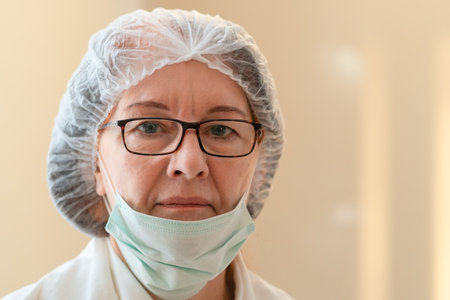 Portrait of a calm and composed female doctor wearing a cap and lowered mask. Her expression conveys confidence and commitment to health and medical care in any condition.の写真素材