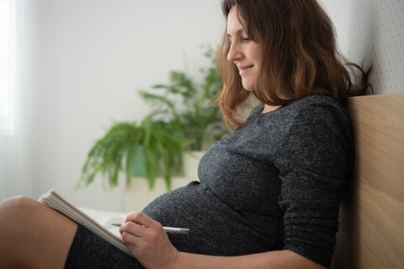 Pregnant woman on bed writing in her notebook. Peaceful time organizing thoughts before birth.