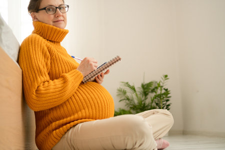 Glowing pregnant woman writing must-have items list for maternity ward in her notebook. Cozy at-home moment of late pregnancy organization.の写真素材
