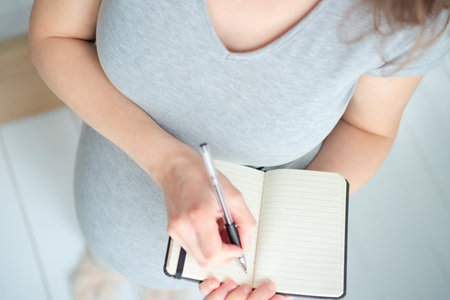 Overhead close-up of expecting mother carefully preparing birth plan in notebook. Organized maternity preparation and important reminders for upcoming delivery.の写真素材