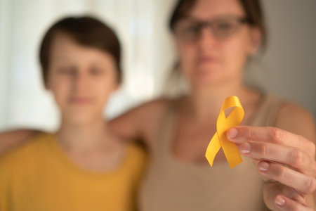 Emotional moment of mother hugging her teenage son who holds yellow satin ribbon symbolizing pediatric cancer awareness and hope.の写真素材