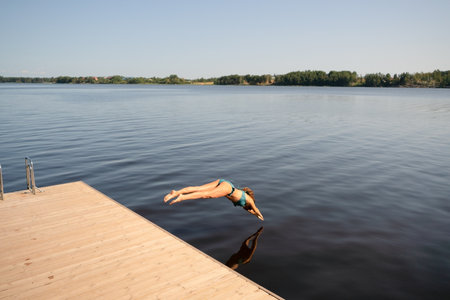 Woman in swimsuit dives into the lake from a pier on a sunny day. Refreshing summer moment full of...