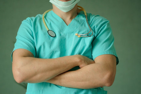 Close-up of a male doctor wearing protective mask and stethoscope, standing with crossed arms....