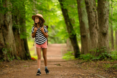 Young girl with a backpack walks forward on a forest path, observing her surroundings with wonder. A calm and curious summer hike during vacation in nature.の写真素材