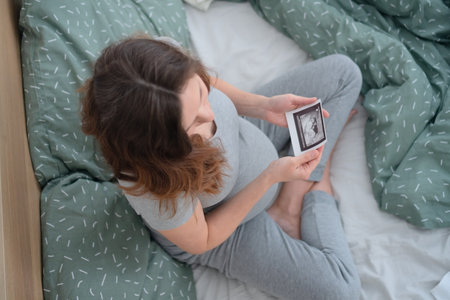 Top-down angle captures pregnant woman cross-legged on bed, lovingly gazing at ultrasound image....