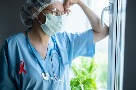A weary female doctor with red awareness ribbon on uniform plans HIV AIDS treatment, showing medical staff hard work and solidarity.の写真素材