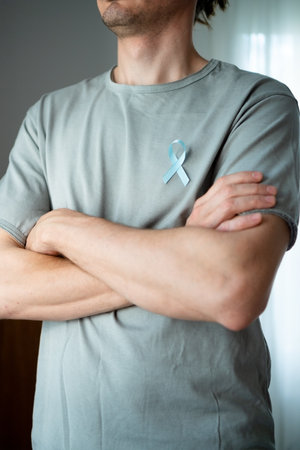 Close up of man with crossed arms and blue satin ribbon on shirt. Symbol of November prostate cancer awareness, early diagnosis, medical help, and support from family and loved ones.の写真素材