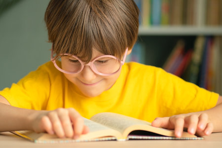 Curious happy child with glasses sits at desk, leaning on hand and looking at open book. Concept of reading, childhood study, learning, early education, preparation for school and knowledge.の写真素材