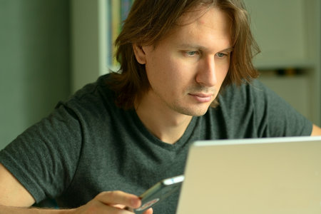 A young man with long hair sits at his desk working intently on a laptop. His gaze is focused on the computer monitor. Home office and remote employment concept.の写真素材
